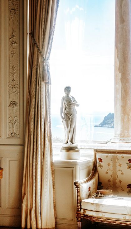 Un salon ancien aux moulures dorées, une statue devant la fenêtre ouverte sur la mer